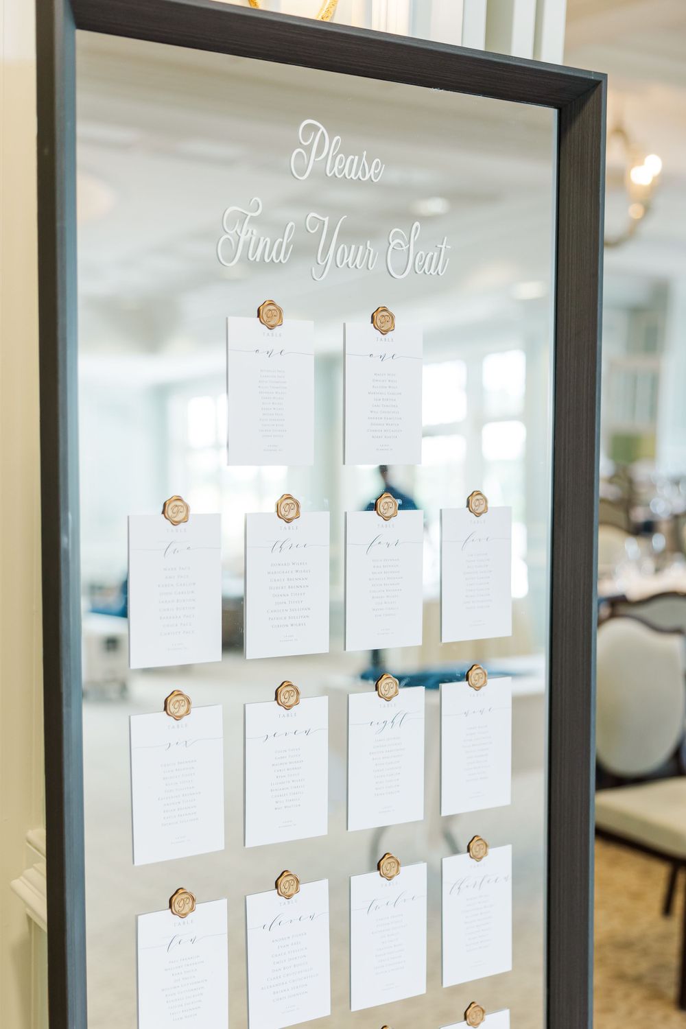 Elegant wedding seating chart display with white cards pinned to a framed board with gold accents and script lettering.