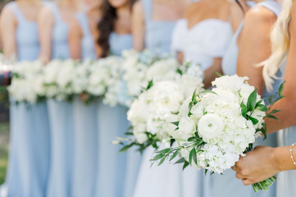 Bridesmaids in light blue dresses hold elegant white hydrangea and eucalyptus bouquets at an outdoor wedding ceremony.