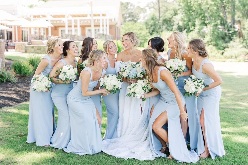 Bridesmaids in flowing light blue dresses holding white bouquets share a joyful moment on a sunny lawn.