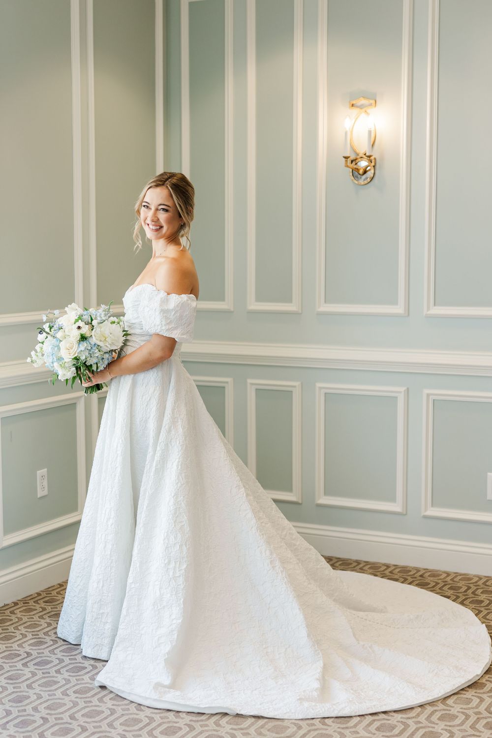 An elegant off-shoulder wedding gown is displayed in a classic room with wainscoting and wall sconces.