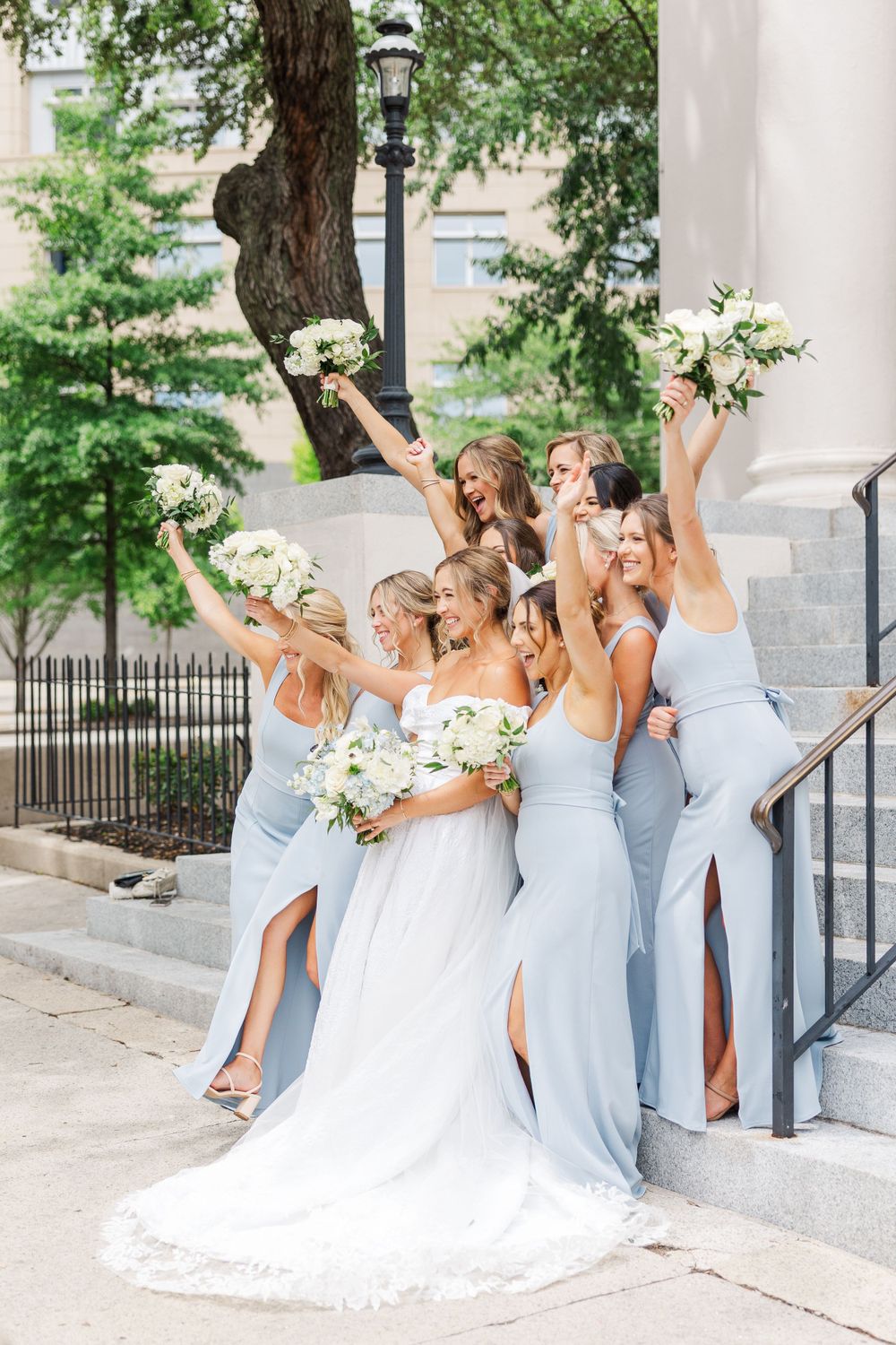 Bridesmaids in light blue dresses and bride in white gown hold bouquets while posing joyfully on church steps.