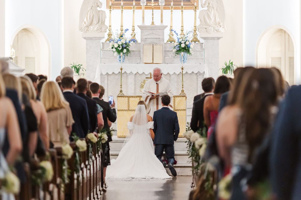 Wedding ceremony taking place in an elegant white church with floral decorations along the pews.