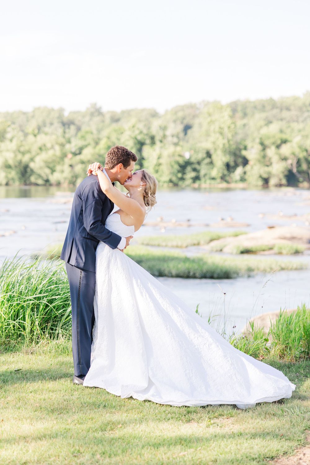 A romantic wedding photo of a couple embracing by a serene lakeside with lush greenery in the background.