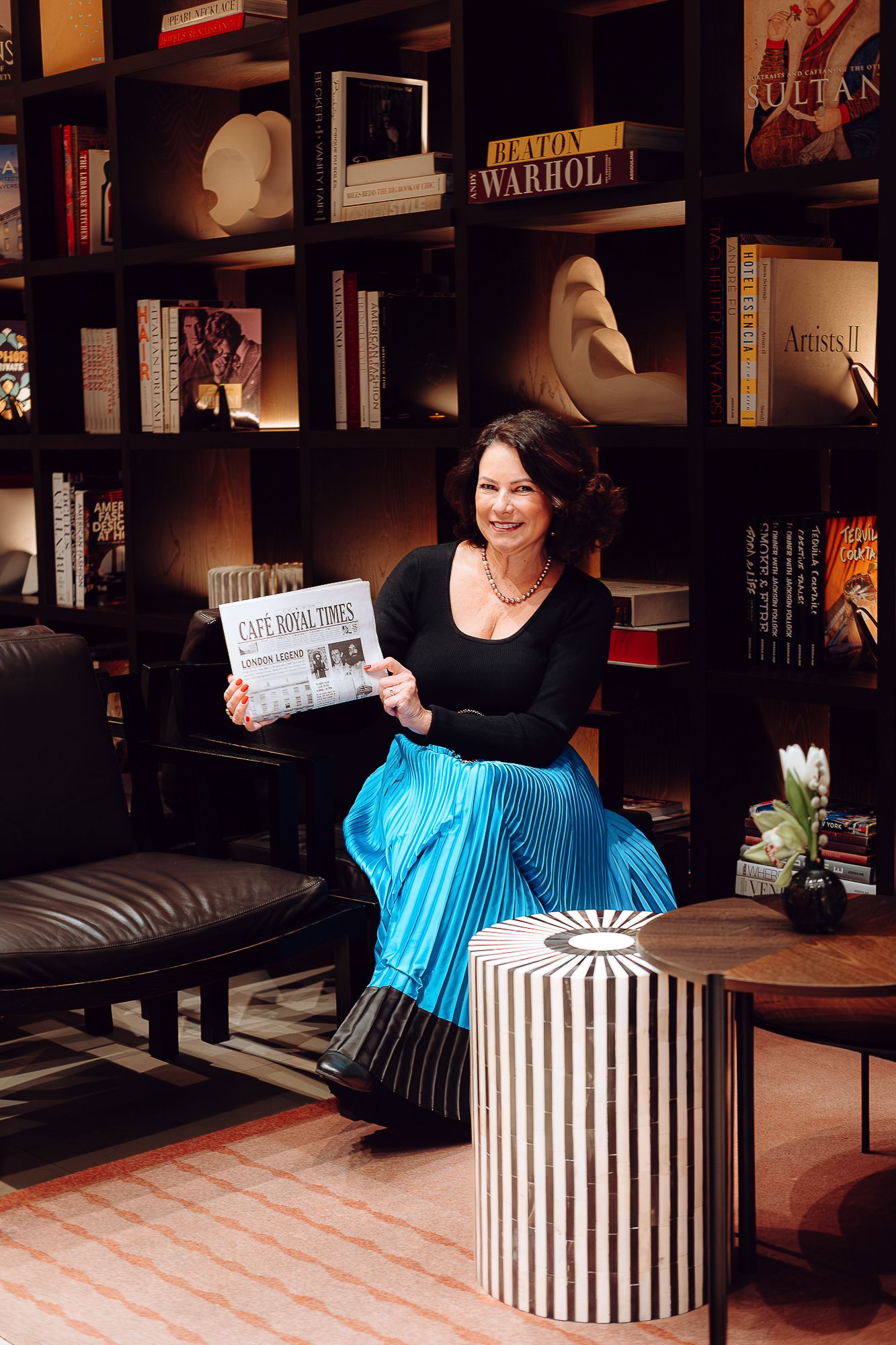 Someone reads against a backdrop of bookshelves while sitting on a leather sofa, wearing a black top and bright blue skirt.
