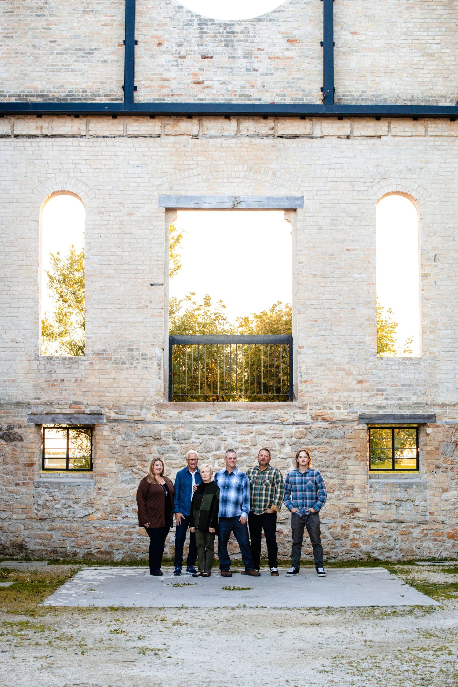 Group stands in front of a rustic stone wall with tall arched windows and metal railings at sunset.