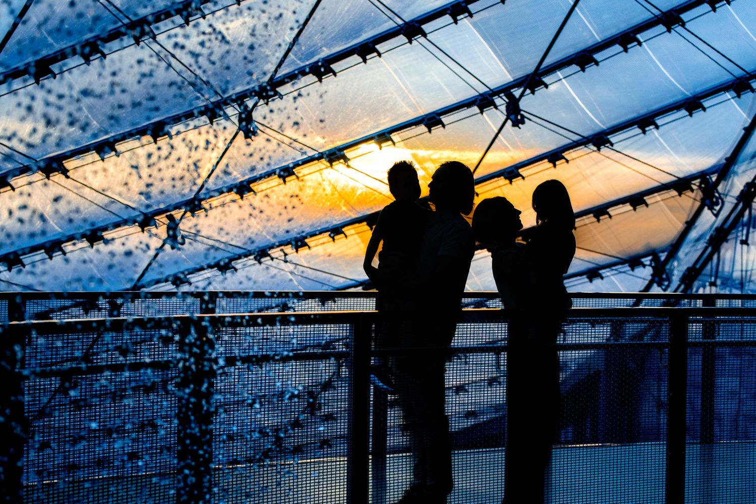 Silhouetted figures standing behind a safety rail at sunset with geometric wire patterns overhead.