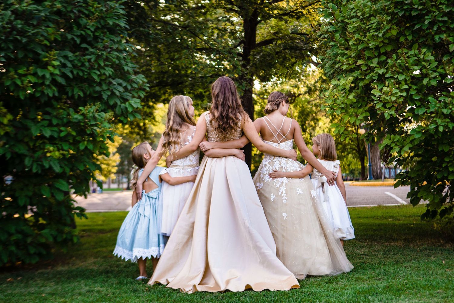 A group of people in formal gowns standing together in a circle with their backs turned in a garden setting.