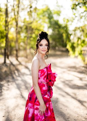 Elegant model in a vibrant pink floral dress poses on a sunlit dirt path surrounded by trees.