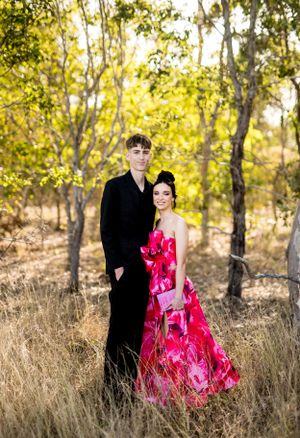 Elegant couple in black suit and pink floral gown pose in golden field at sunset.