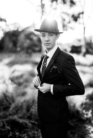 Black and white formal portrait featuring classic suit, tie and fedora in natural outdoor setting.