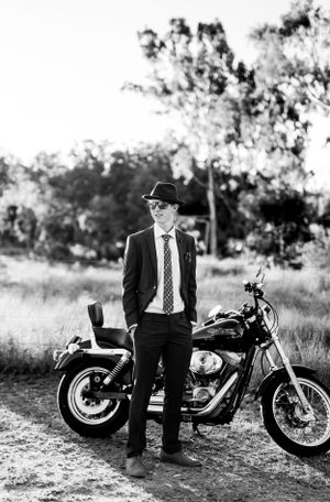 Black and white portrait showing classic suit and fedora next to vintage motorcycle in outdoor setting.