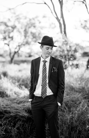 Black and white portrait of suited figure with fedora hat in rustic outdoor setting.