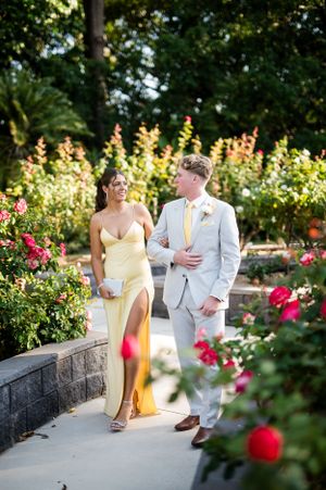 A couple in yellow and grey formal attire walks along a garden path lined with blooming flowers.