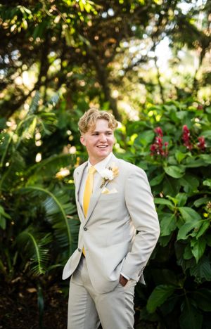 A person in a light grey suit and yellow tie smiles while standing in a tropical garden setting.