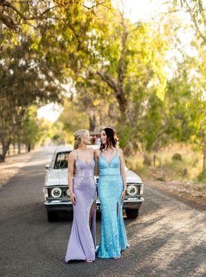 Two evening gowns in blue and lavender shimmer against a vintage car backdrop.
