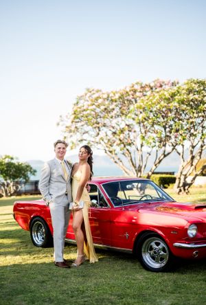A dressed-up couple poses with a classic red Ford Mustang on a sunny lawn.