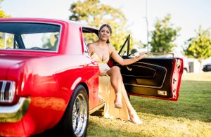 A person in a yellow dress poses playfully in the door of a classic red Mustang.