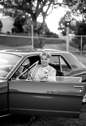 A black and white portrait of someone sitting in the driver's seat of a classic car.