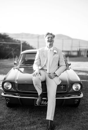 A classic Ford Mustang with someone in a light suit sitting on the hood in a black and white photograph.