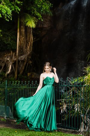 An elegant green gown flows in the breeze against an ornate iron fence and waterfall backdrop.