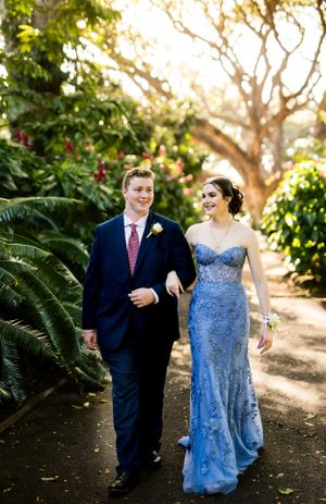 Couple walking arm in arm through garden path in formal blue gown and navy suit.