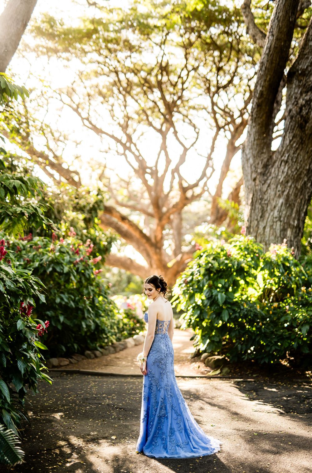 Long blue formal dress photographed from behind on a scenic garden pathway.
