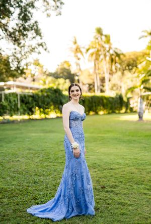 Blue lace formal gown photographed on a sunlit grassy lawn with palm trees.
