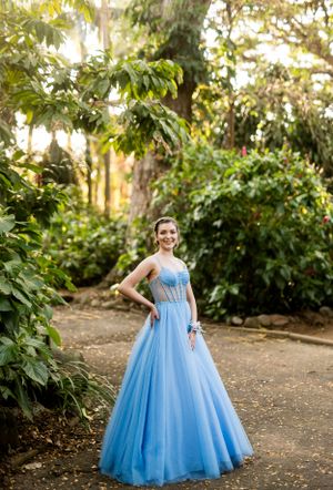 A flowing light blue ball gown photographed in a garden setting with natural sunlight filtering through trees.