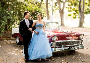 A formal couple poses in front of a vintage burgundy Chevrolet on a tree-lined path.
