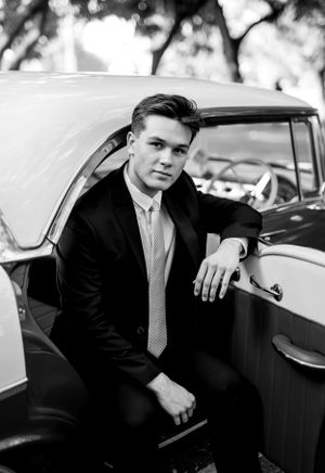 Black and white portrait of a young man in formal attire leaning against a vintage car.