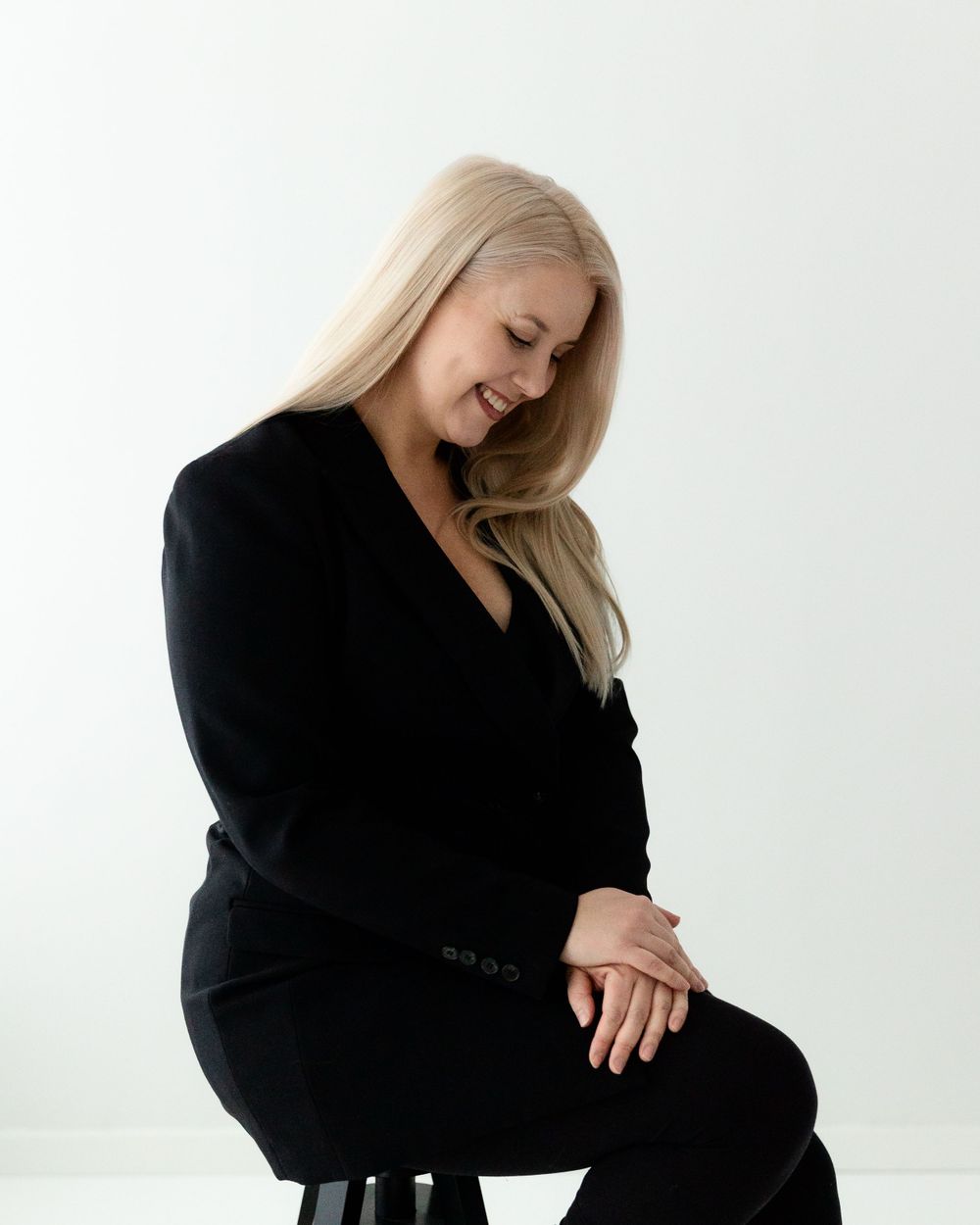 A woman in a black outfit sits elegantly on a stool against a white backdrop in a minimalist studio setting.