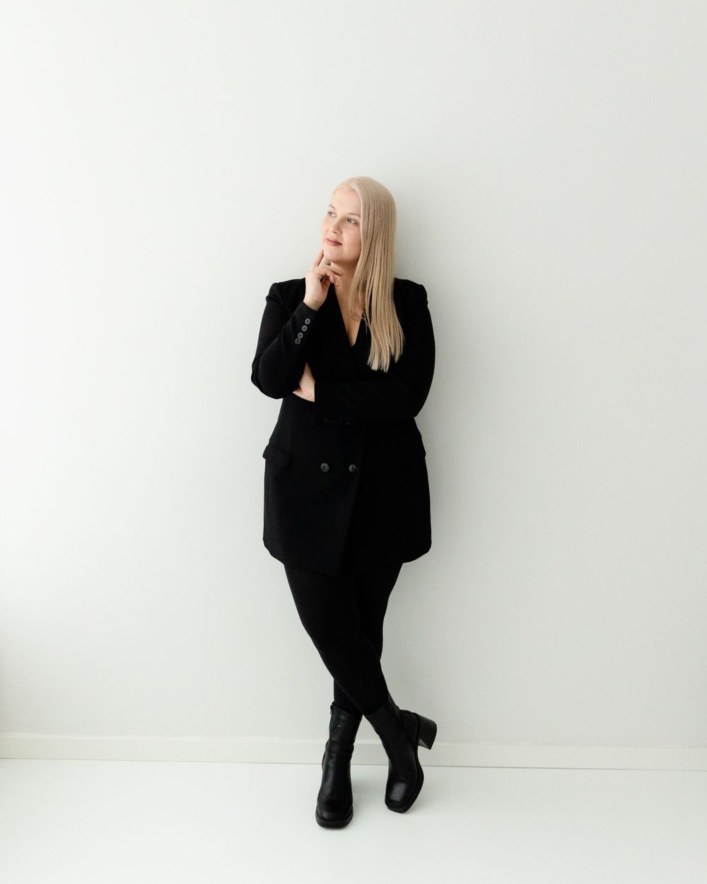 A person in all black attire poses thoughtfully against a white wall, wearing a long blazer, pants and ankle boots.