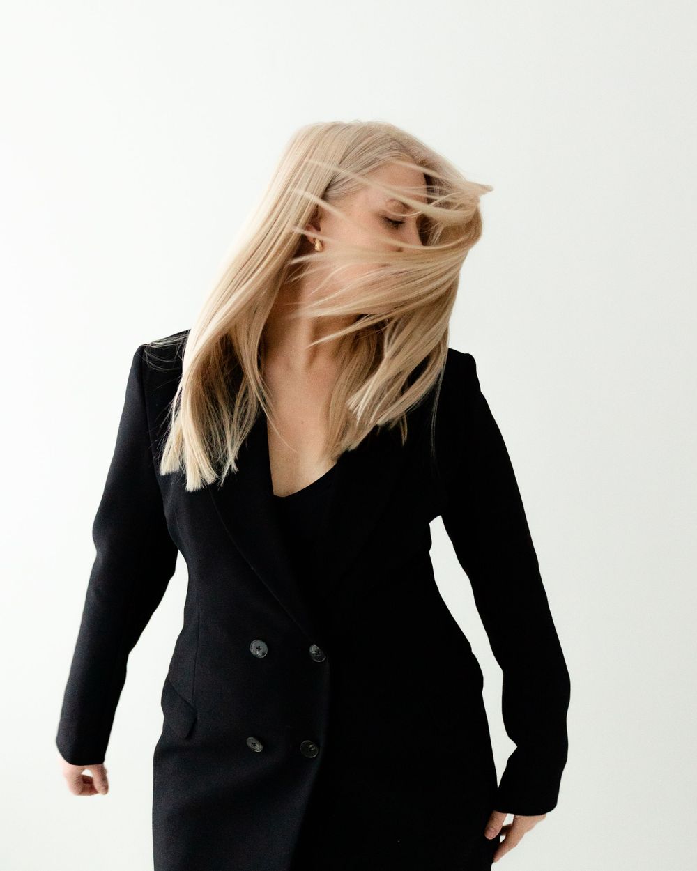 Blonde hair flips dynamically against white backdrop as model wears classic black double-breasted blazer dress.