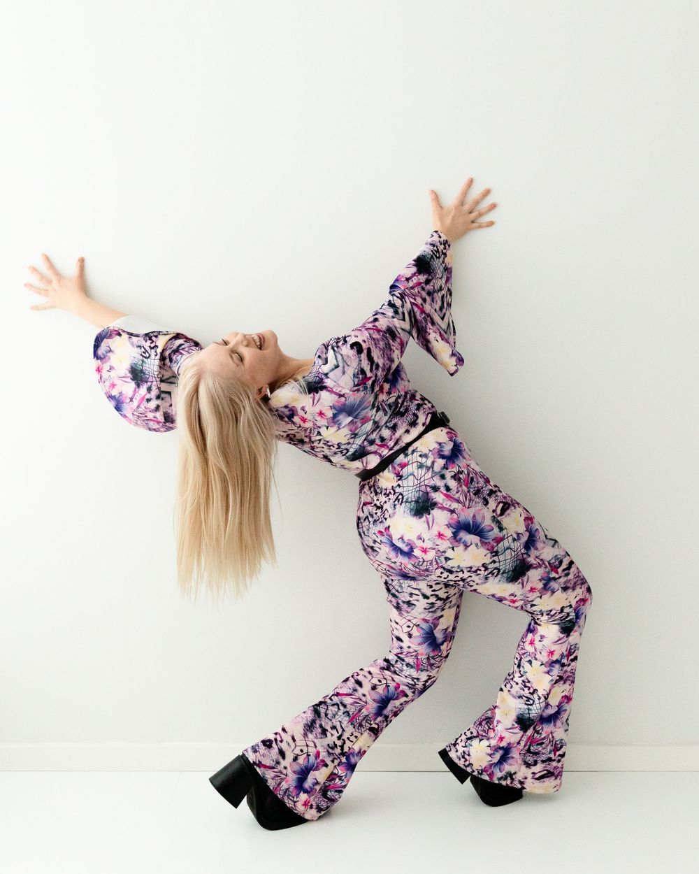 Dynamic pose of performer in purple floral jumpsuit with arms outstretched against white wall.