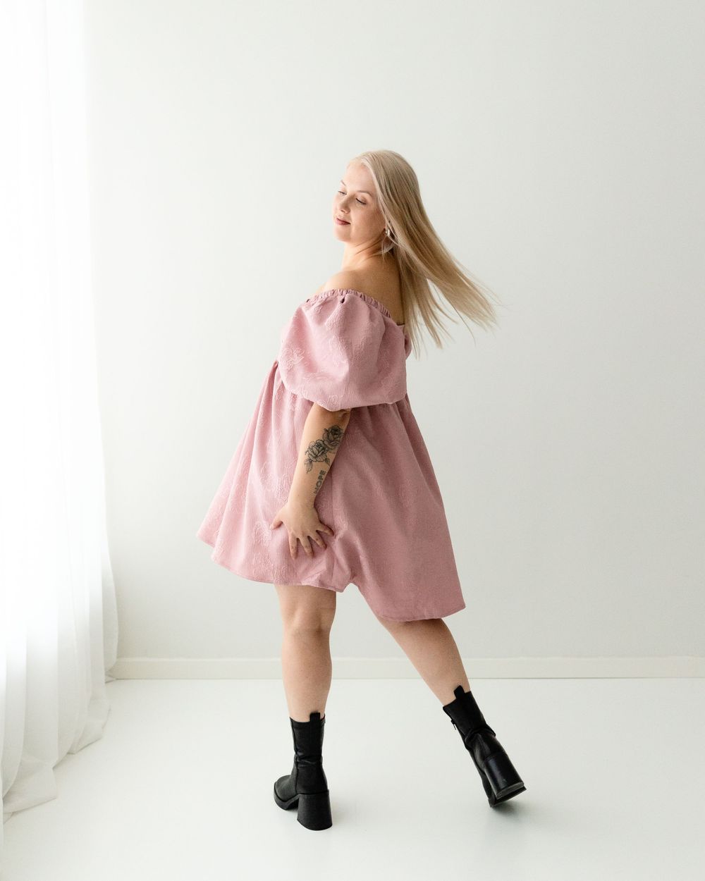 A woman in a flowing pink off-shoulder dress and black boots poses dynamically against a white background.