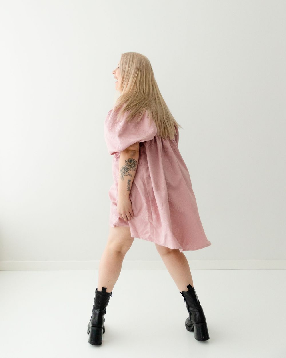 Person with long blonde hair twirling in pink dress and black boots against white background.
