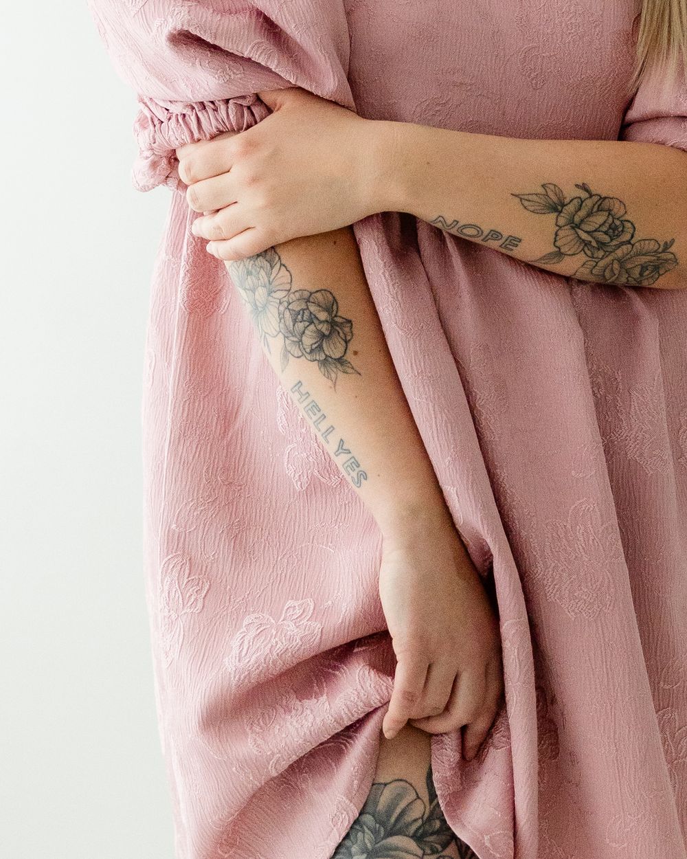 Arms with floral tattoos rest against soft pink fabric showing delicate black ink botanical designs.