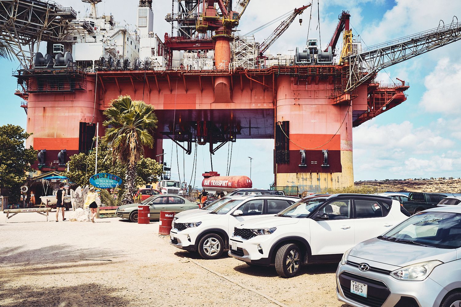 Parked cars beneath large red industrial crane at waterfront shipping facility.