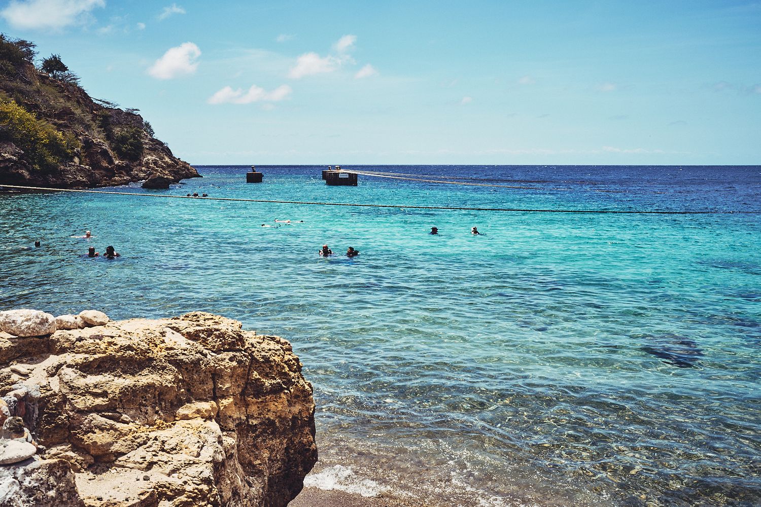 Swimmers enjoying crystal clear turquoise waters in a rocky beach cove with boats in the distance.