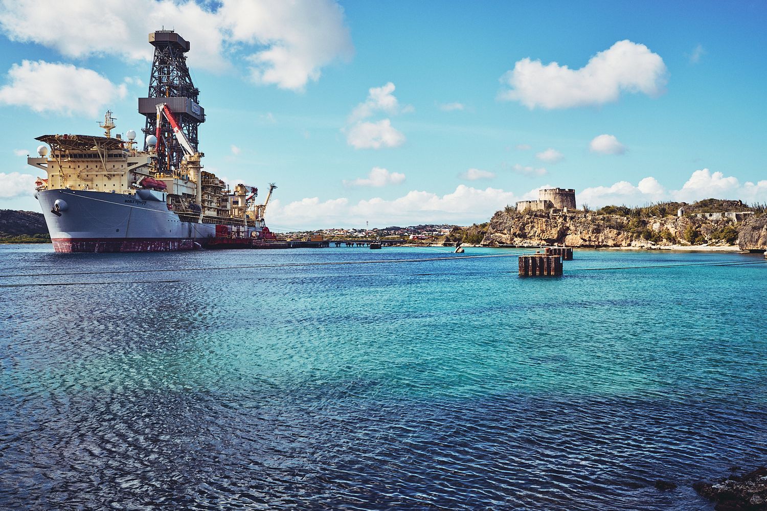 Massive offshore drilling vessel in bright turquoise waters near coastal cliffs.