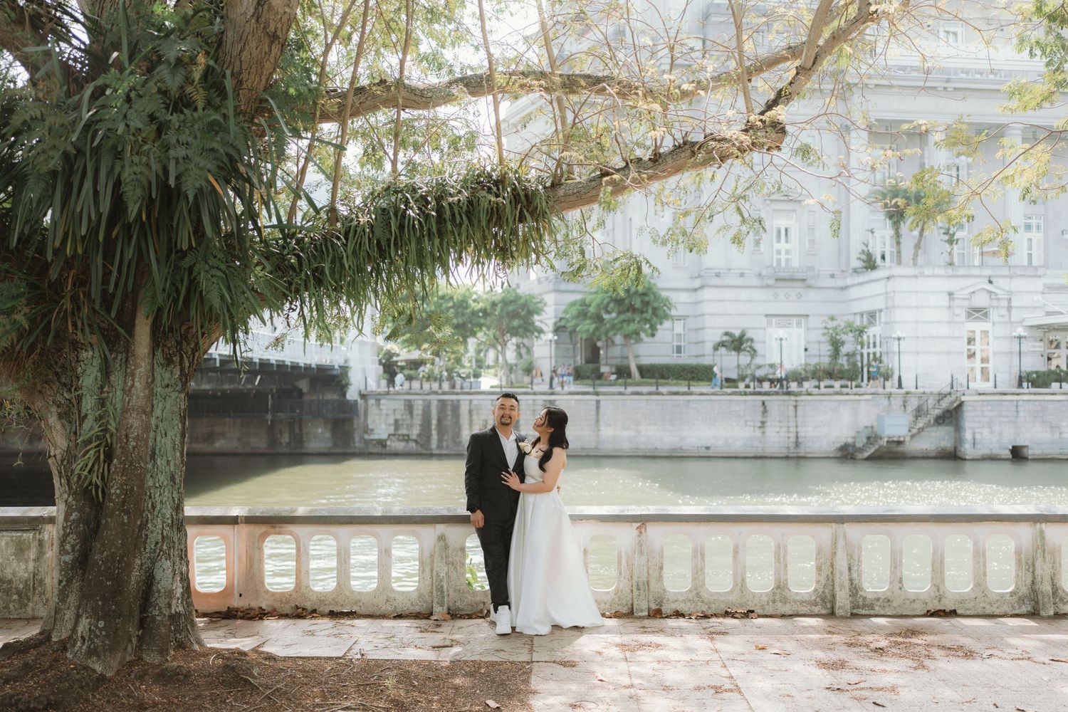 A couple in wedding attire stands beneath a large tree on a stone terrace overlooking a historic building.