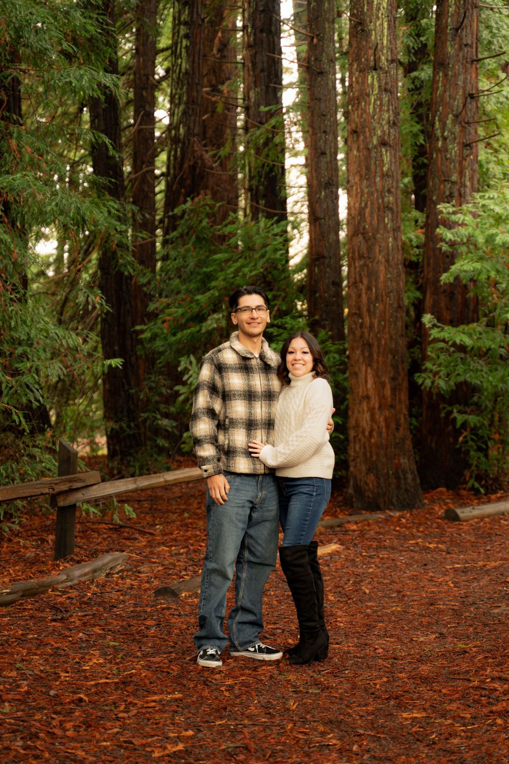 A couple shares intimate moments among tall redwood trees.