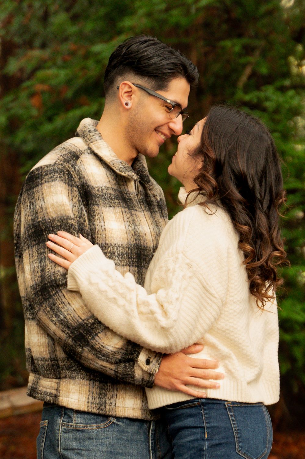 A couple in cozy sweaters embraces in a redwood forest setting.