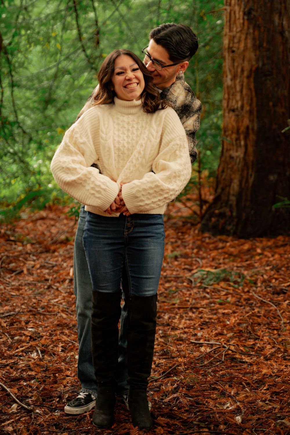 Couple in casual fall attire embracing in a forest setting with autumn leaves on the ground.