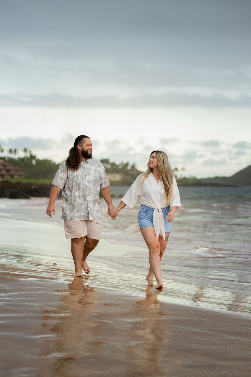 Couple walking hand in hand on a beach at sunset wearing casual summer outfits.