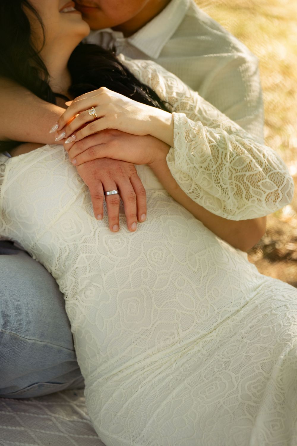 Detail shot of hands with wedding rings against white lace dress and denim in golden sunlight.