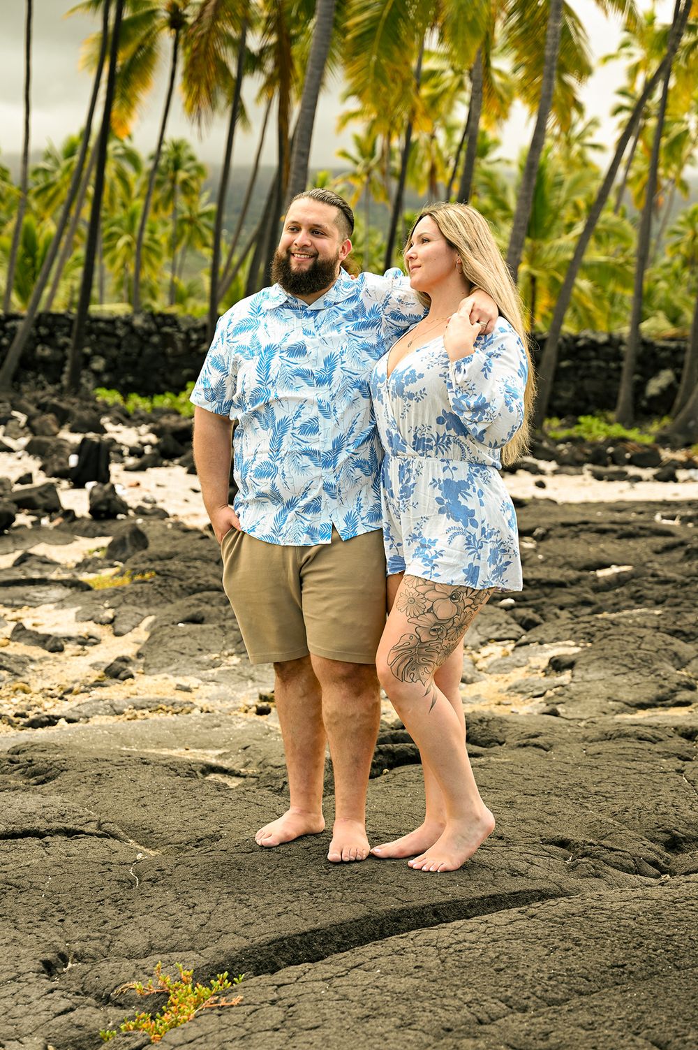 A couple wearing matching tropical prints poses on volcanic rocks with palm trees.