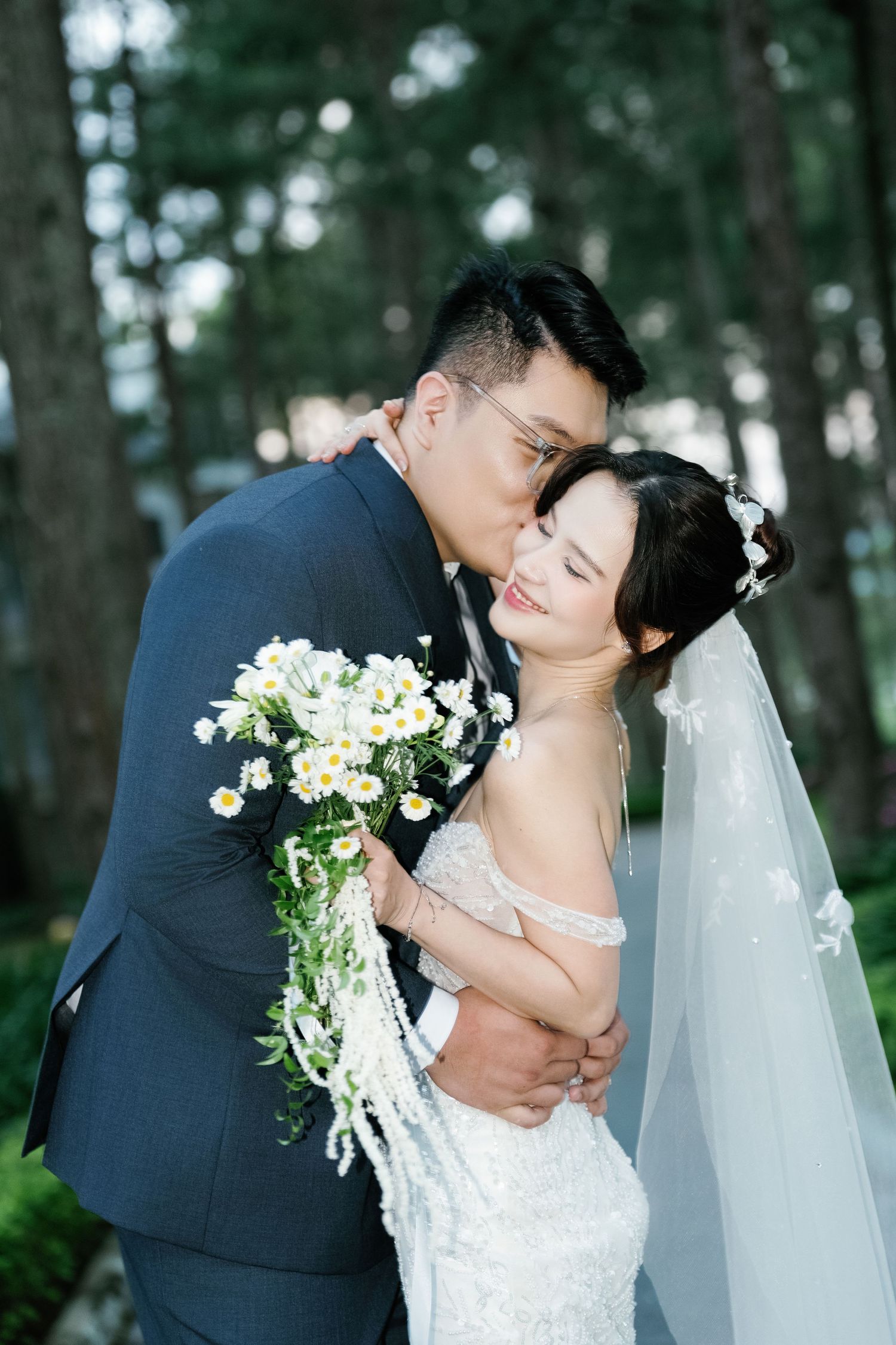 Newlyweds share a tender embrace in a forest setting, with the bride holding a white floral bouquet and wearing a flowing veil.
