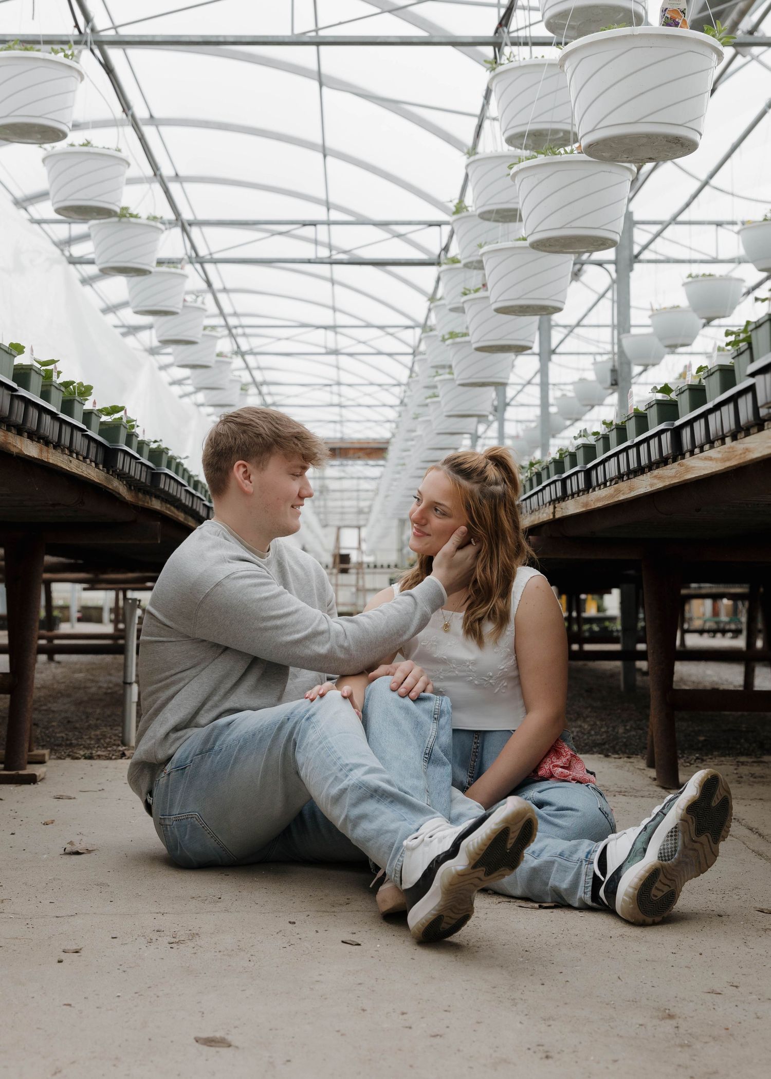 A young couple sits intimately on the floor of a greenhouse with white ceiling panels and rows of plants.