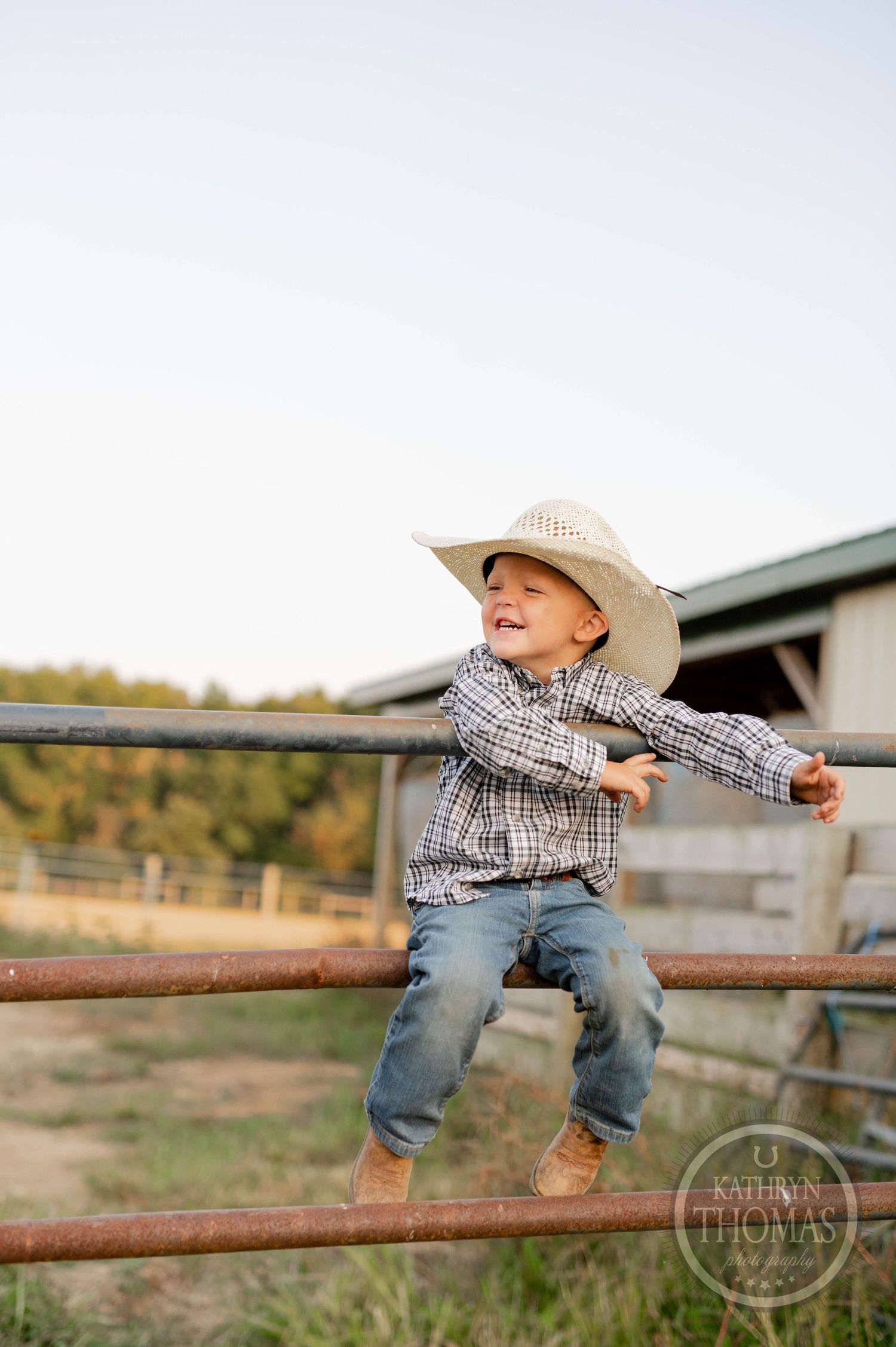 Young cowboy in plaid shirt and straw hat balancing on metal fence rail at ranch.